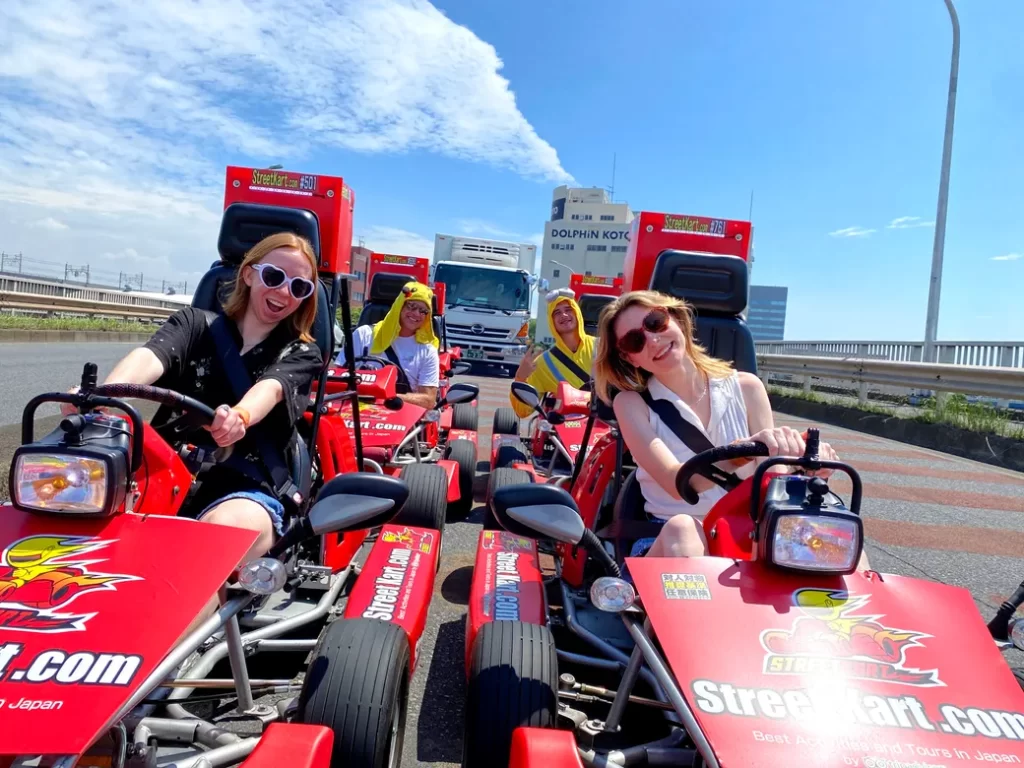 Group of friends wearing sunglasses ride red Street Kart go‑karts on a sunny road, smiling and racing side by side near trucks in the background.